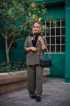 Fashionable woman in suit and headscarf posing outdoors with handbag.