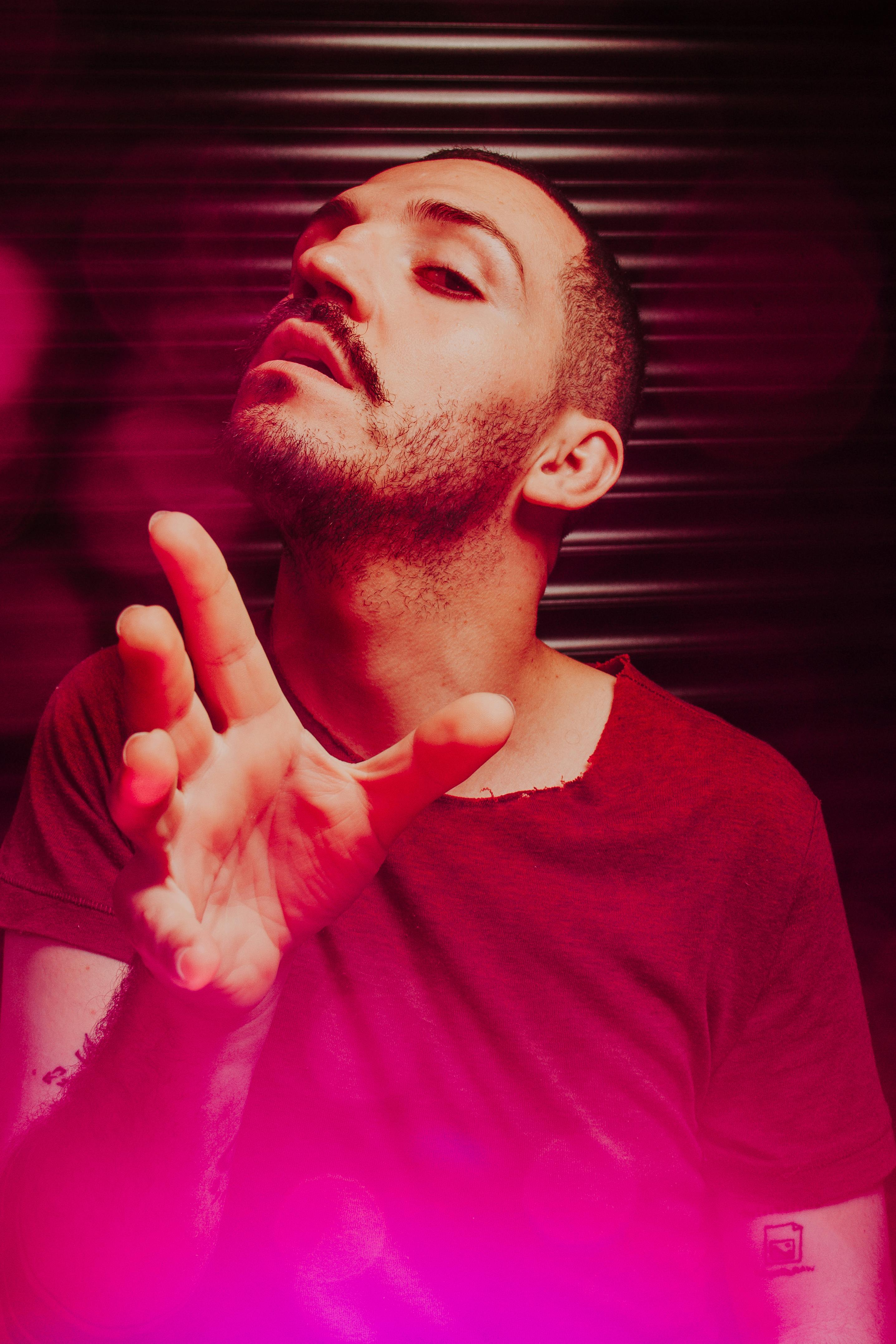 Dynamic portrait of a man with a mustache in a red shirt reaching forward, featuring artistic blur effect and pink lens flare.