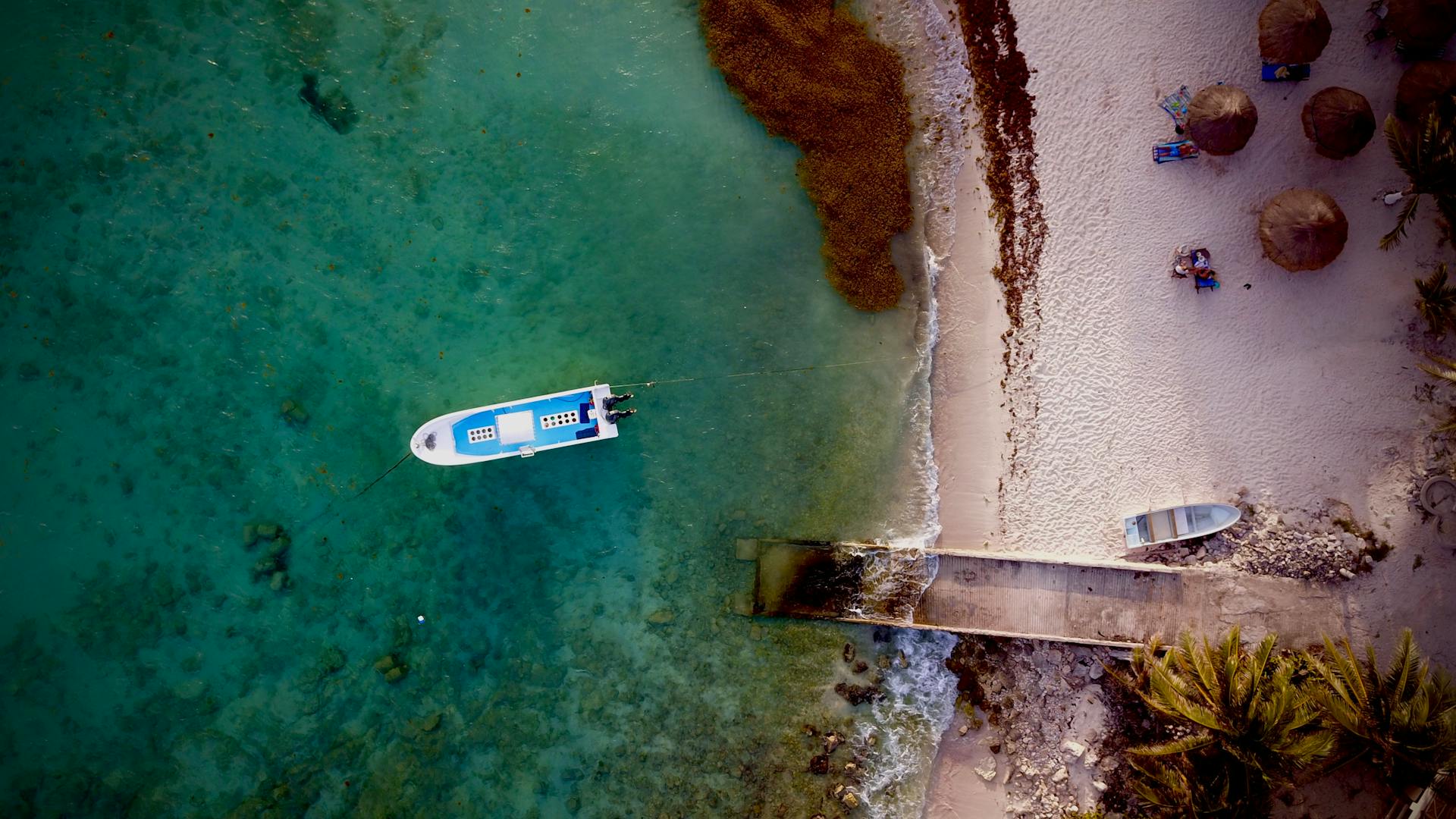 Aerial photo of a turquoise sea, moored boat, and sandy shore in Paa Mul, México.