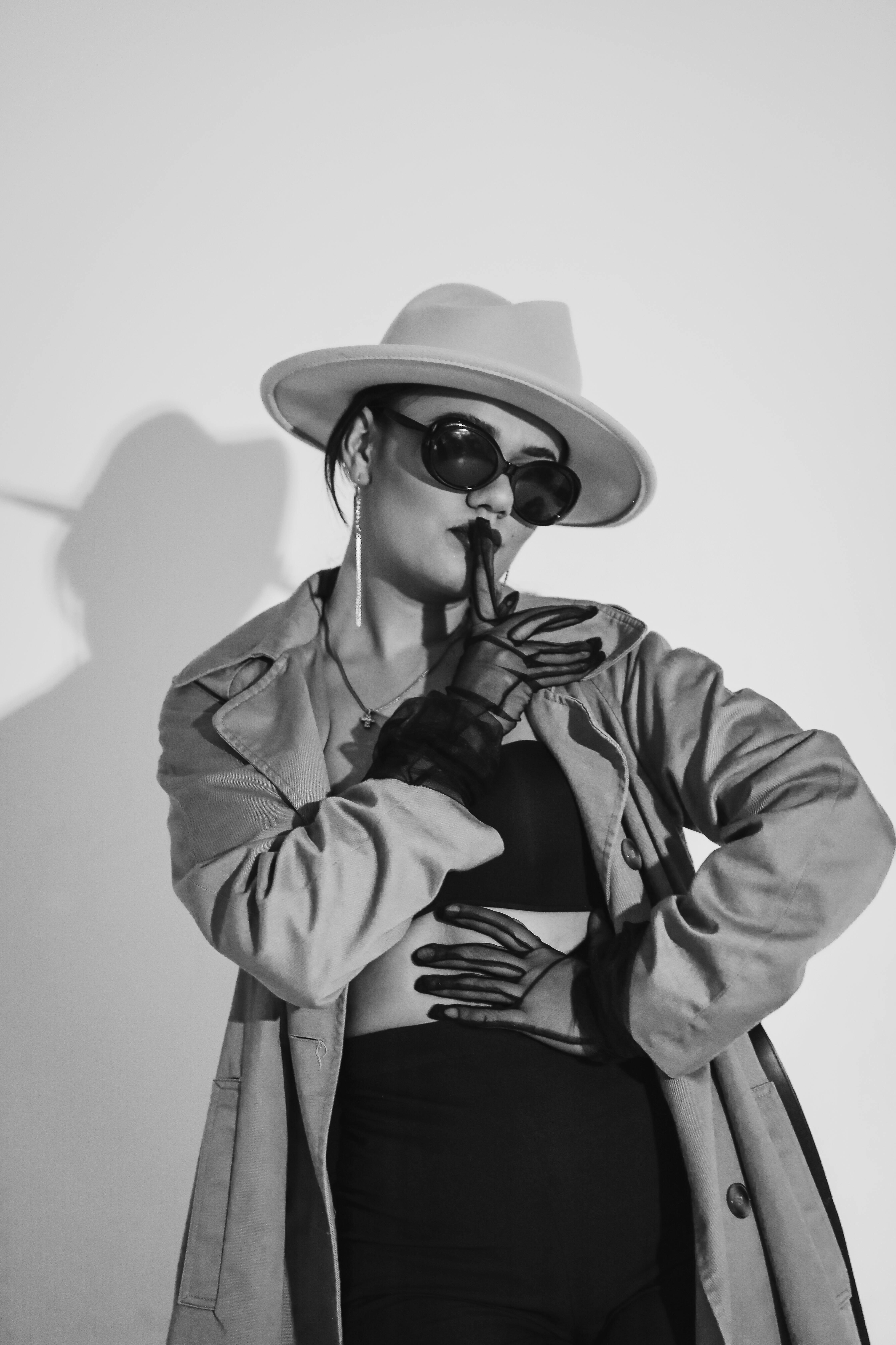 Free Black and white fashion photo of a woman in a trench coat, hat, and sunglasses posing with confidence. Stock Photo