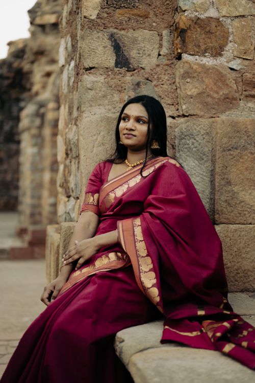 Free A woman in a traditional red sari poses gracefully against an ancient stone wall, embodying elegance and cultural attire. Stock Photo