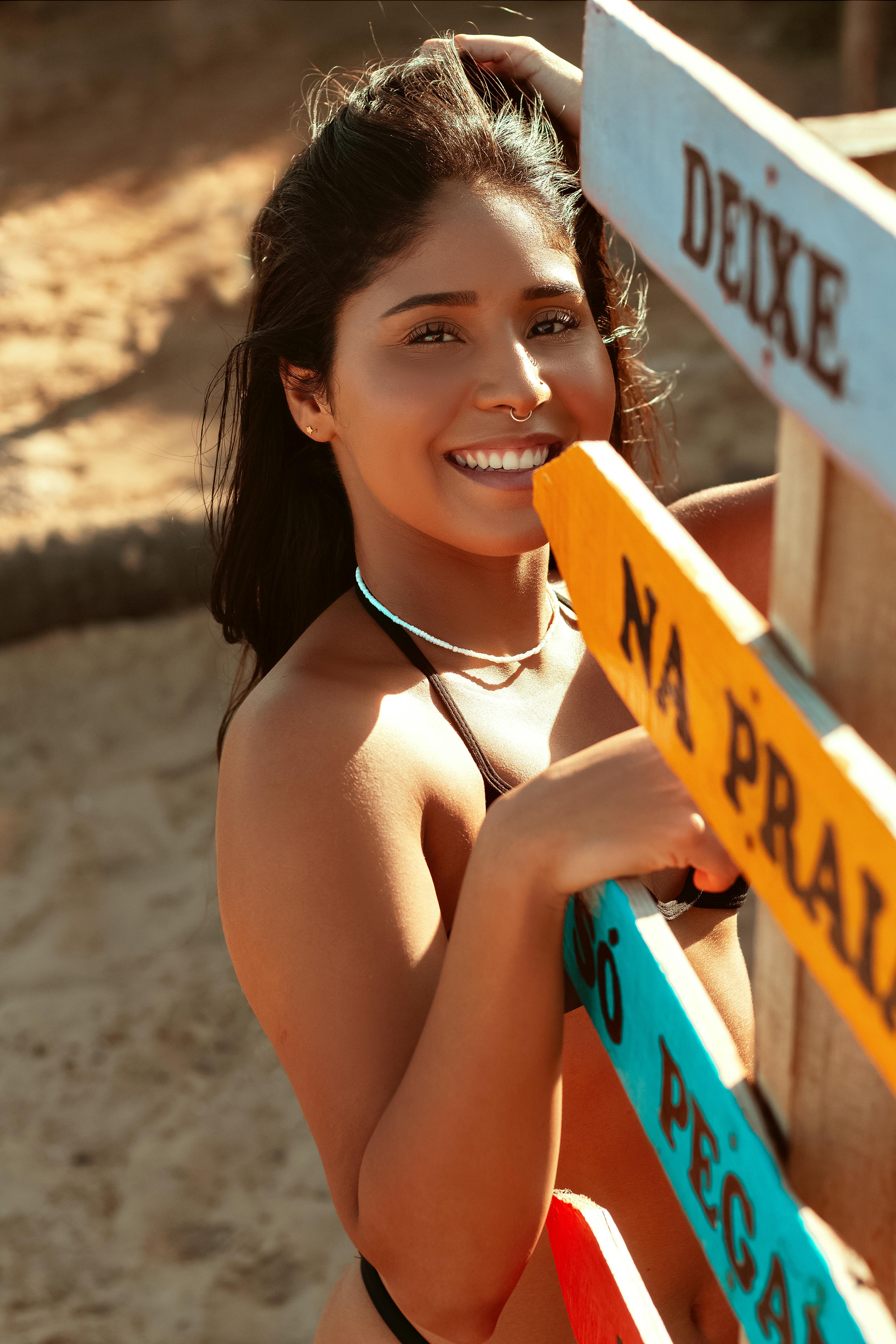 A young woman smiling beside vibrant signs at the beach, enjoying the summer sun.