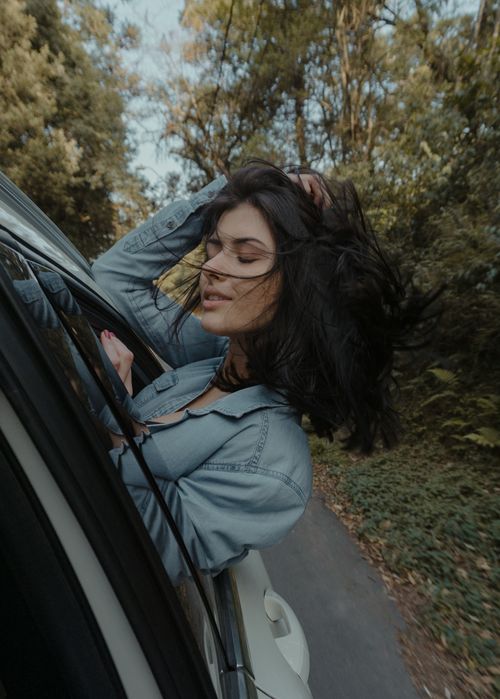 Free Young woman leaning out of a car window enjoying the wind in a scenic forest drive. Stock Photo