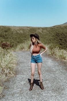 Fashionable woman in hat and shorts poses confidently on a scenic country road.
