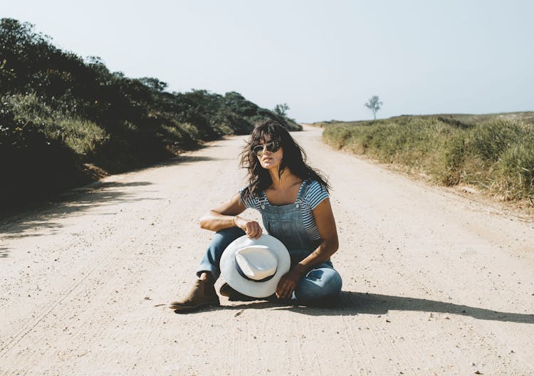 Woman Wearing Denim Overalls And Sunglasses Sitting On The Road In The Countryside 