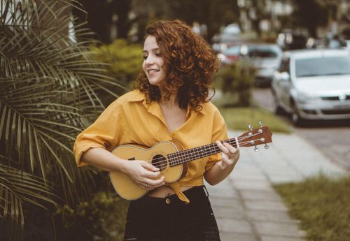 Free A cheerful woman with curly hair plays a ukulele outdoors on a city sidewalk. Stock Photo