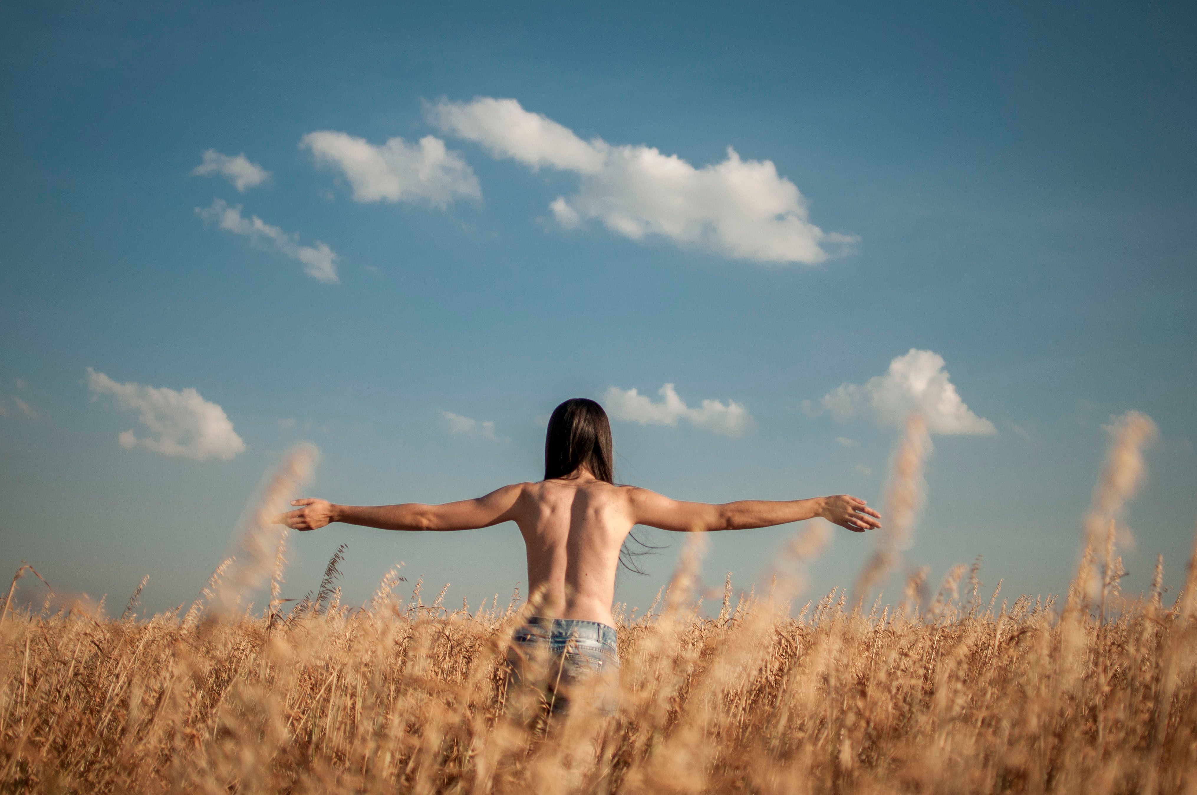 Back view of a shirtless woman with long hair enjoying freedom in a wheat field under a bright blue sky.