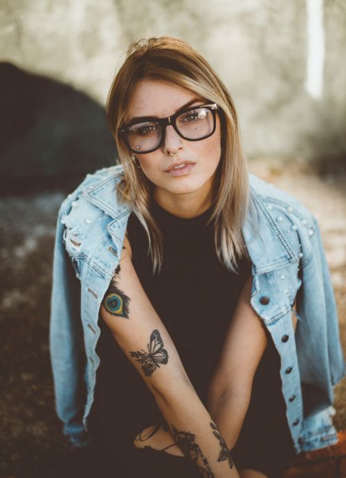 Free Portrait of a stylish young woman with glasses and tattoos wearing a denim jacket outdoors. Stock Photo