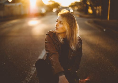 A woman in a leather jacket sits on a sunlit street capturing the golden hour ambiance.