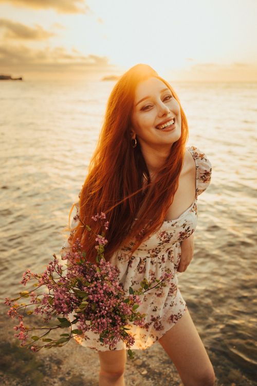 Free Smiling woman with red hair holds flowers by the sea at sunset, exuding warmth and happiness. Stock Photo