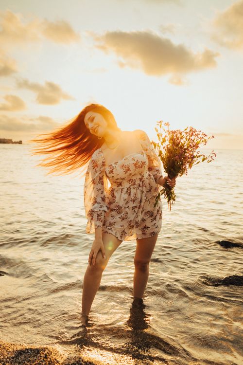 Free A woman in a floral dress stands in the water with a bouquet, enjoying a sunset by the sea. Stock Photo