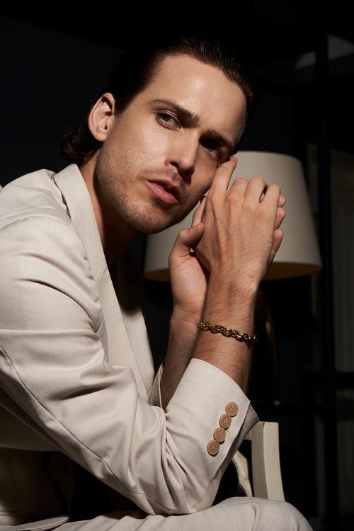 Free Stylish male model in a white suit sitting indoors, offering a thoughtful pose in a modern setting. Stock Photo