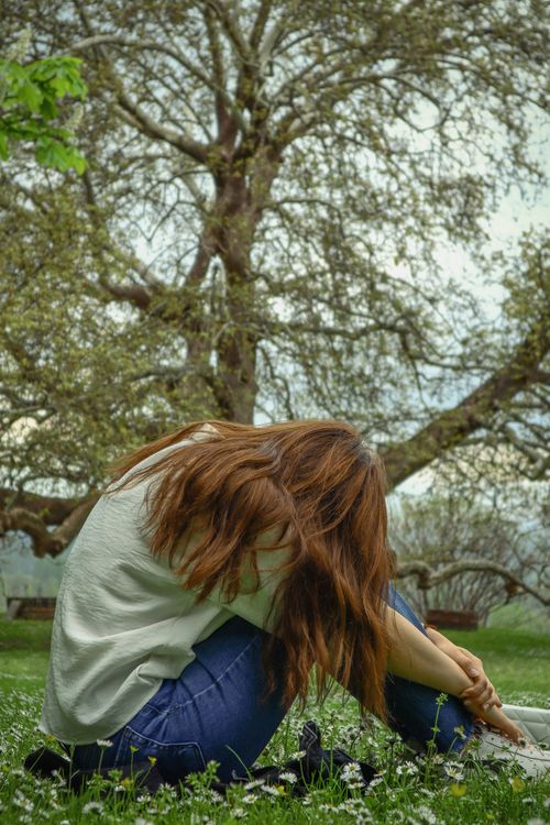 Free Woman with long hair sitting under a tree in a grassy meadow, contemplative pose. Stock Photo