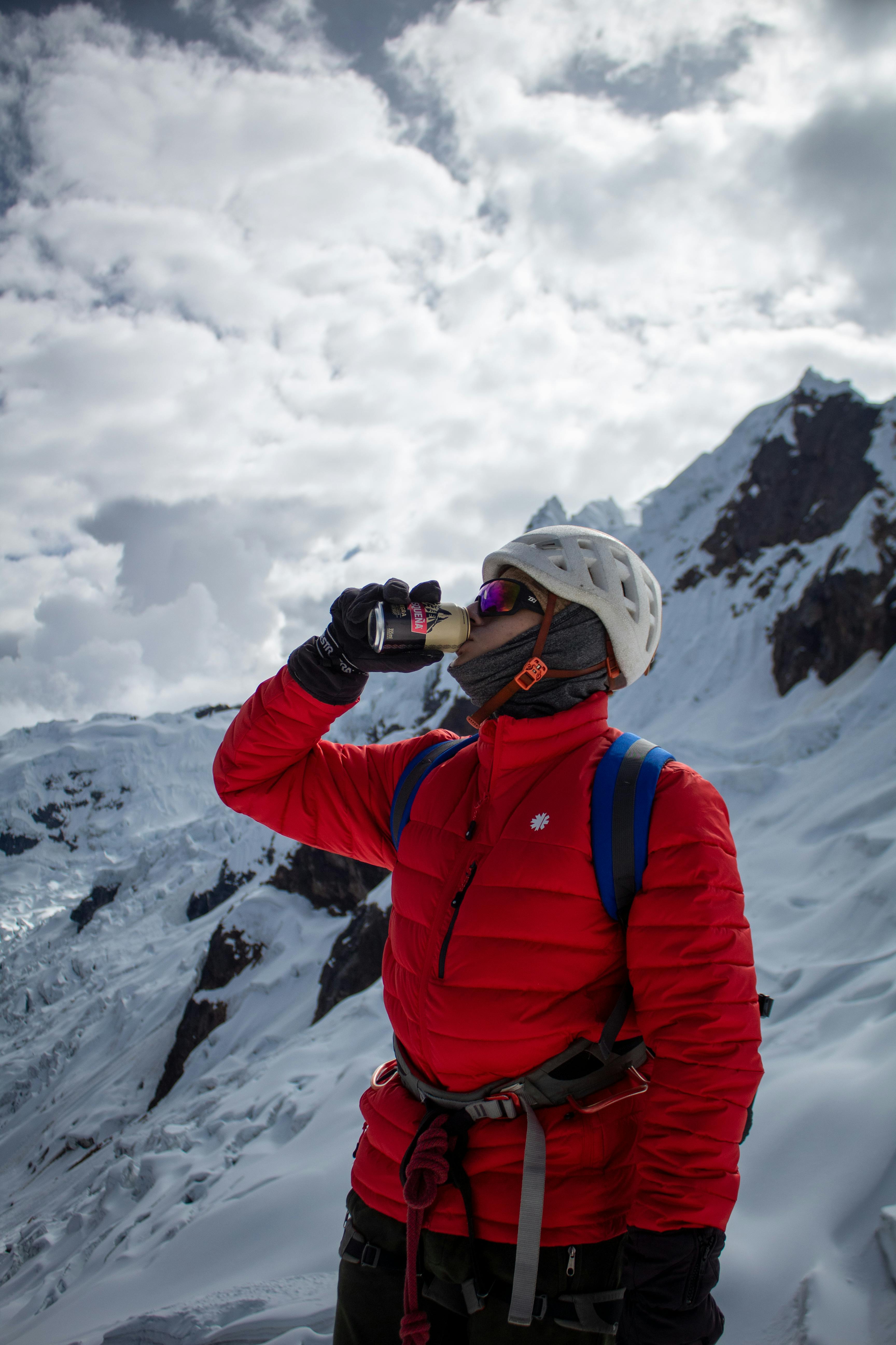 A climber in a red jacket drinks from a can on a snowy mountain peak under cloudy skies.