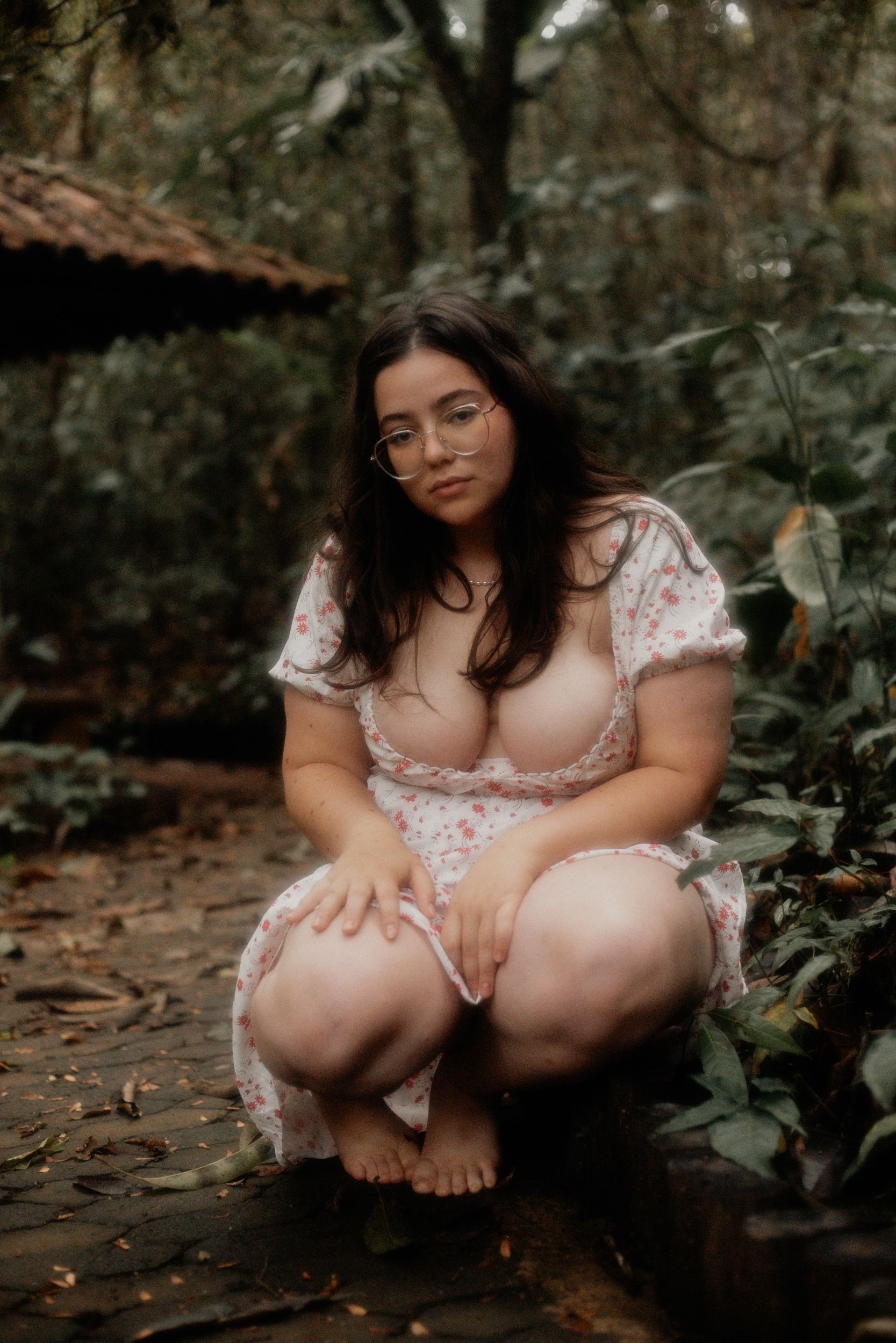 A young woman crouches barefoot in a lush forest in Belo Horizonte, exuding natural beauty.