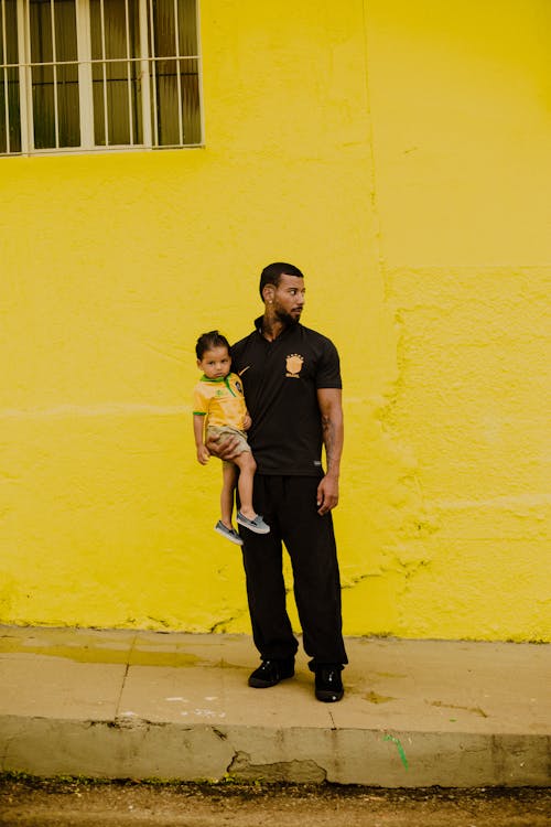 Man Standing on a Sidewalk in front of a Yellow Building and Holding His Little Daughter 