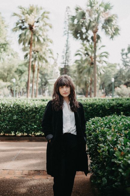 Elegant woman in black blazer and white shirt, standing in a sunny park with palm trees.