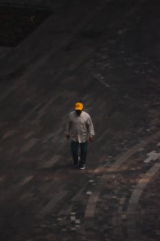 A man in a yellow cap walks alone on a dark paved pathway, captured from above.