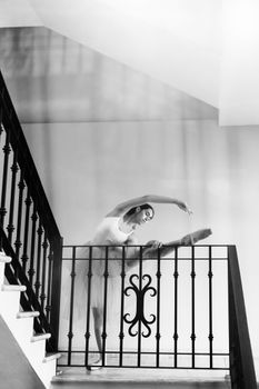 Black and white image of a ballerina stretching on a staircase railing indoors.