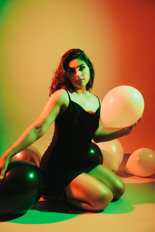 Free Woman in a stylish dress kneeling amidst colorful balloons with a vibrant background. Stock Photo