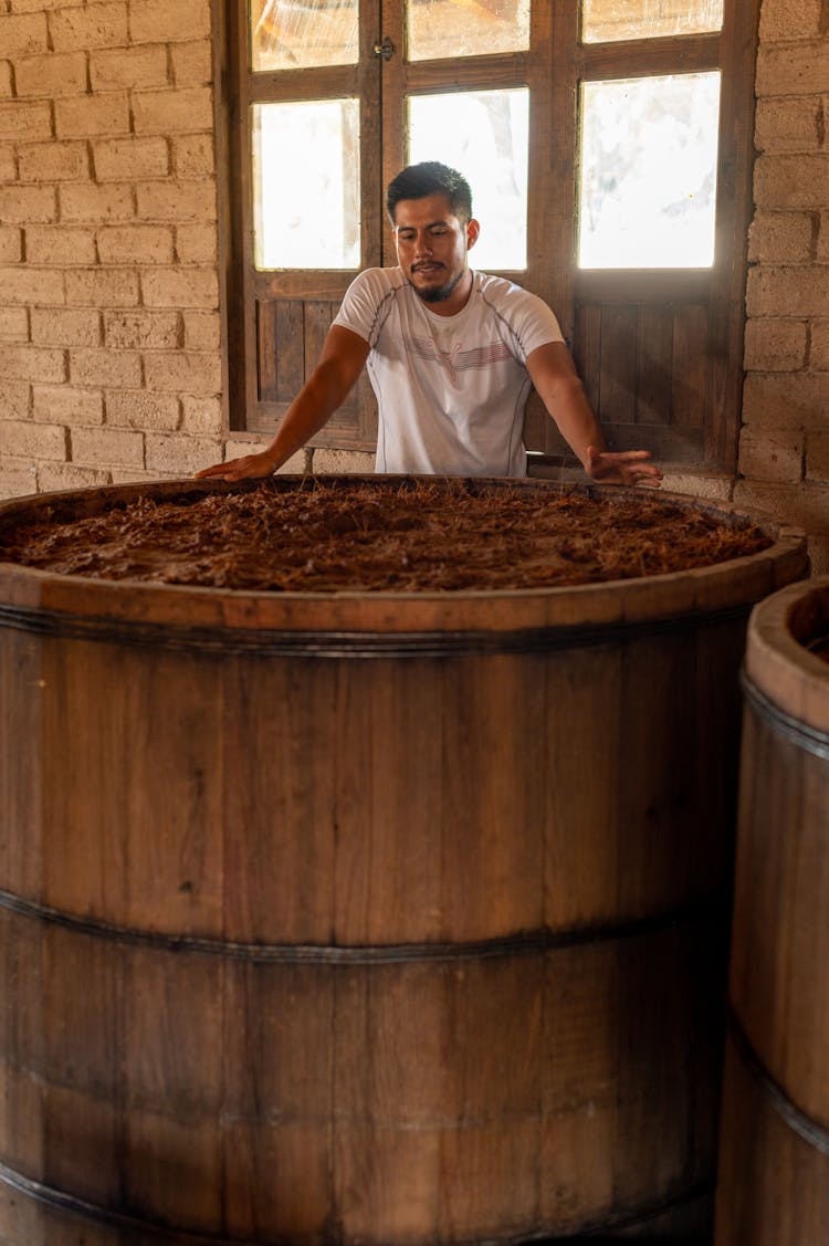 Man Standing Next To A Large Container With Agave At A Mezcal Distillery 