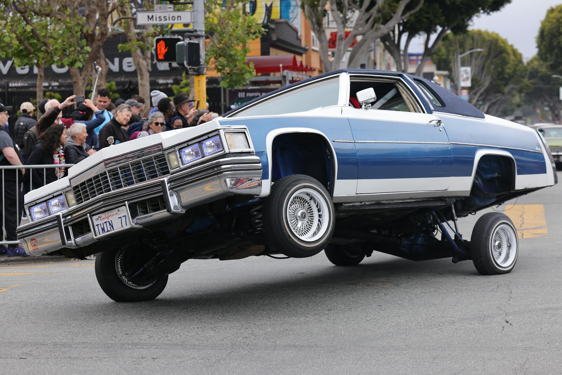 A blue lowrider car performing stunts at a lively urban car show event.