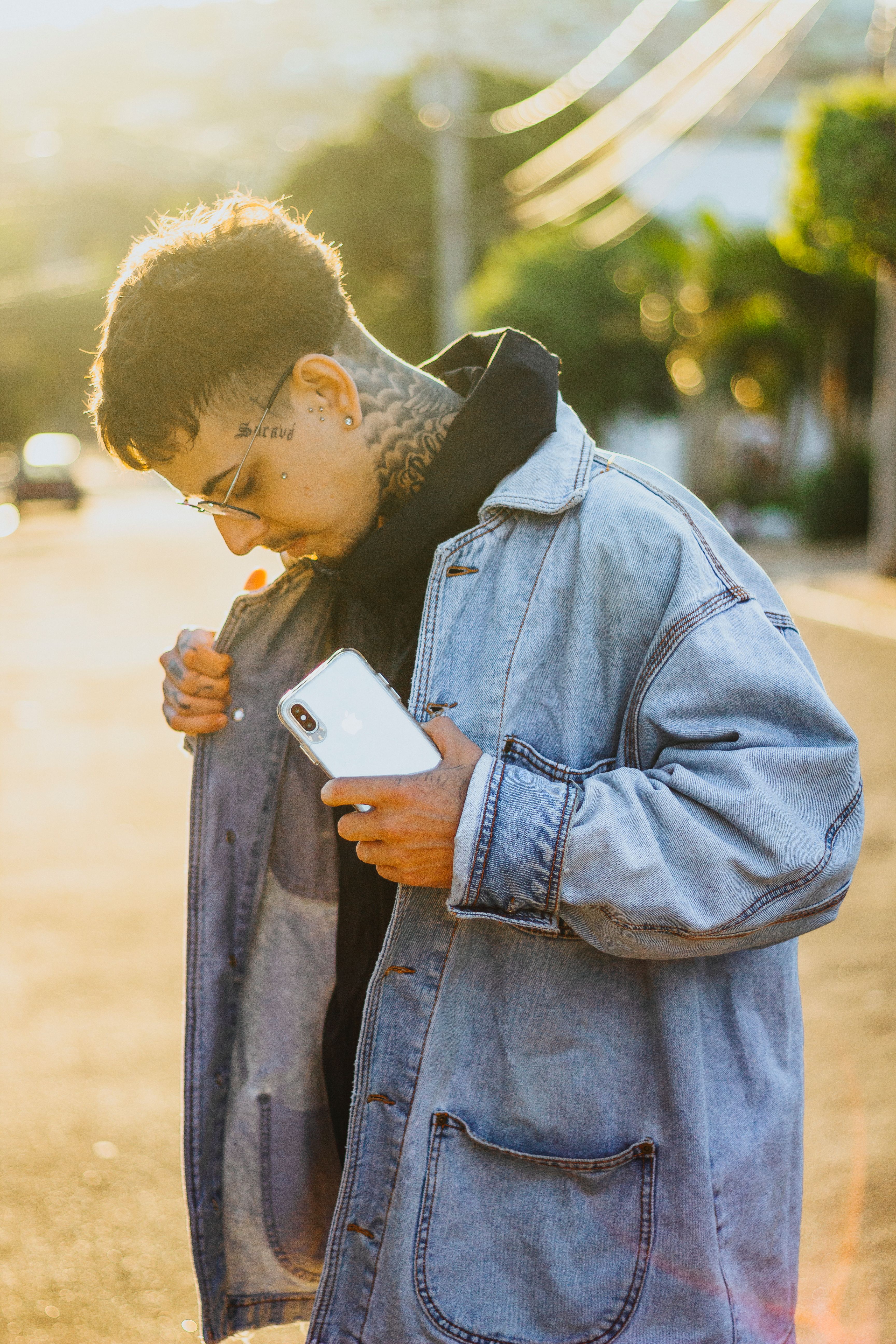 Casual street style portrait of a young man in a denim jacket with a smartphone on a sunny day.