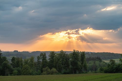 Free Stunning sunset with sunbeams breaking through clouds over lush green fields and trees. Stock Photo
