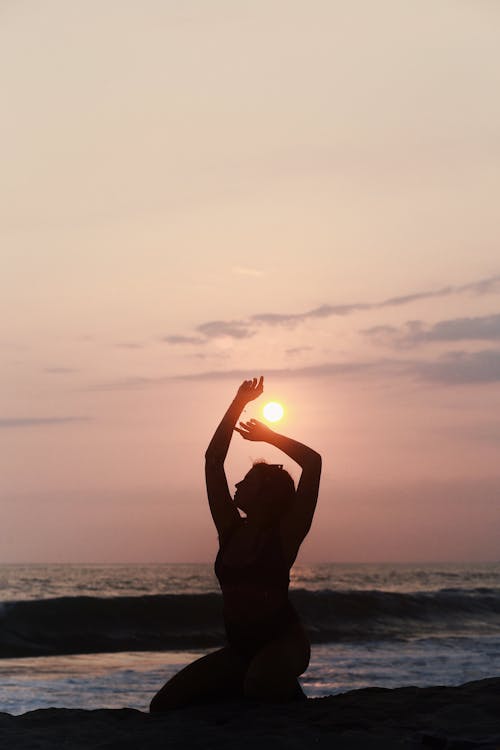 Silhouette of a Woman Sitting on a Beach at Sunset with Her Arms Raised