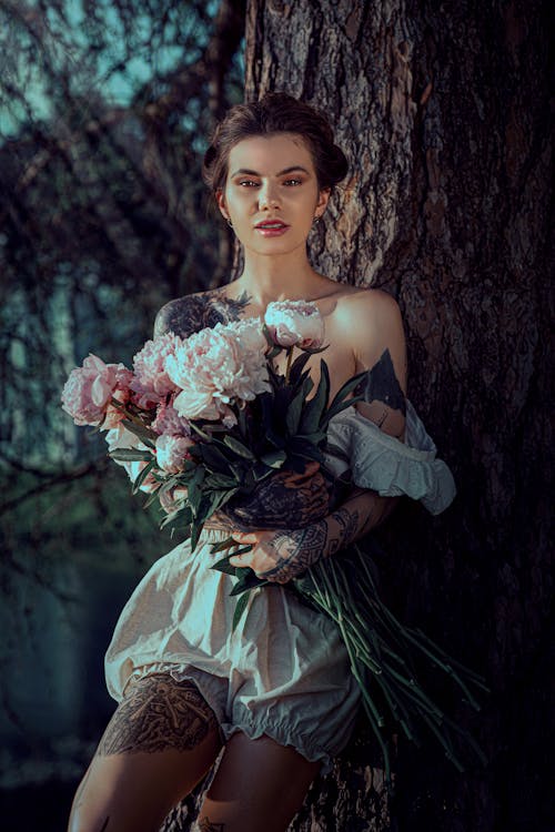Young Woman with Tattoos Standing by a Tree and Holding a Bunch of Flowers