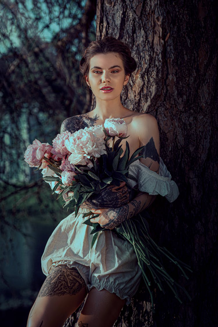 Young Woman With Tattoos Standing By A Tree And Holding A Bunch Of Flowers