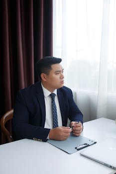 Young professional man in a suit sitting at a desk in an office setting, looking thoughtful.