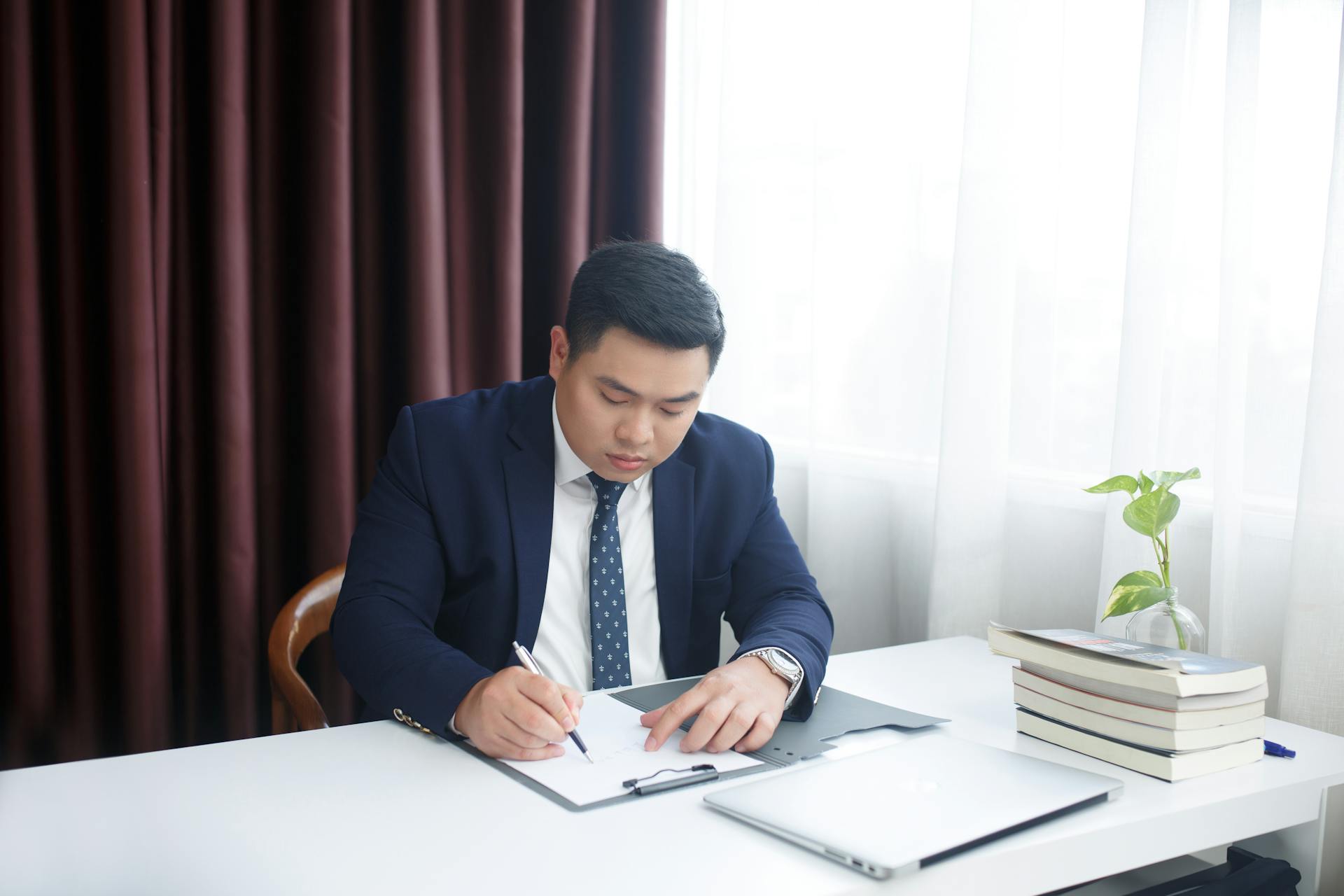 Asian businessman in a suit writing at his desk in an office environment, focusing on documents.