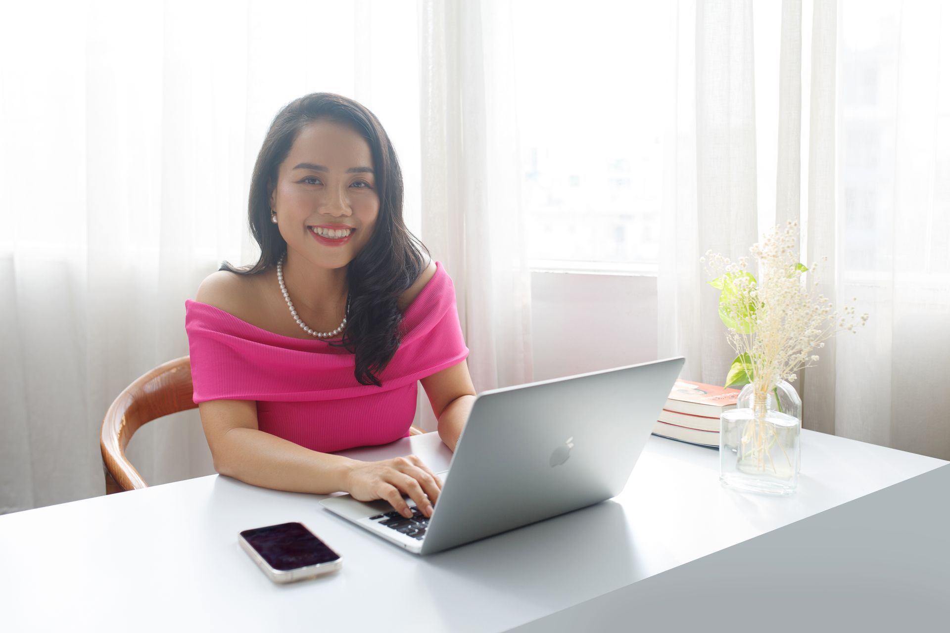 Asian woman smiling while working on a laptop at home office desk.