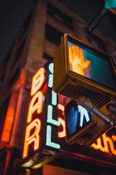 A city street scene featuring a neon bar sign with a pedestrian traffic light at night.