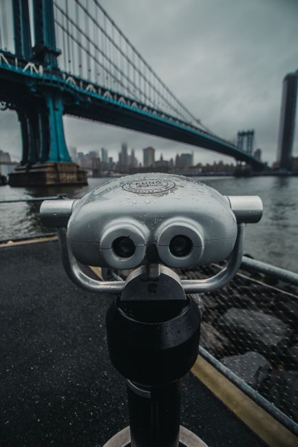 Close-up of a tower viewer with a city skyline and iconic bridge in the background on a rainy day.
