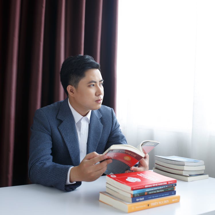 Photo Of A Pensive Young Man In A Suit Sitting At A Table With Books