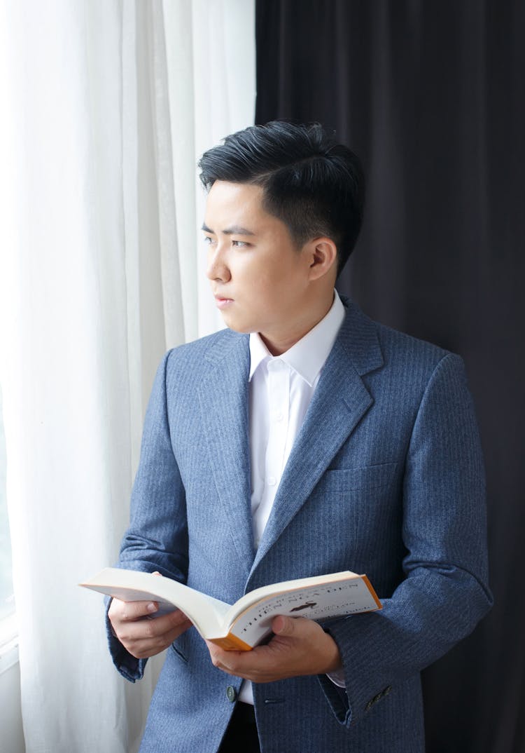 Photo Of A Young Man In A Suit Standing By The Window And Reading A Book