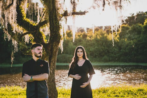 Man Standing with Pregnant Woman under Wisteria Tree on River Bank