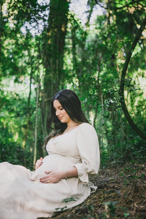 Pregnant Woman in a White Dress Sitting on the Ground in the Forest 