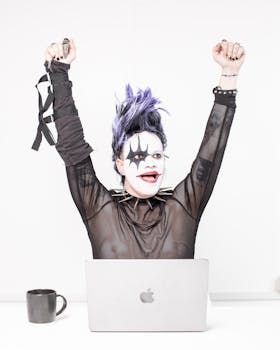 A person in goth makeup and clothing celebrates energetically at a desk with a laptop.