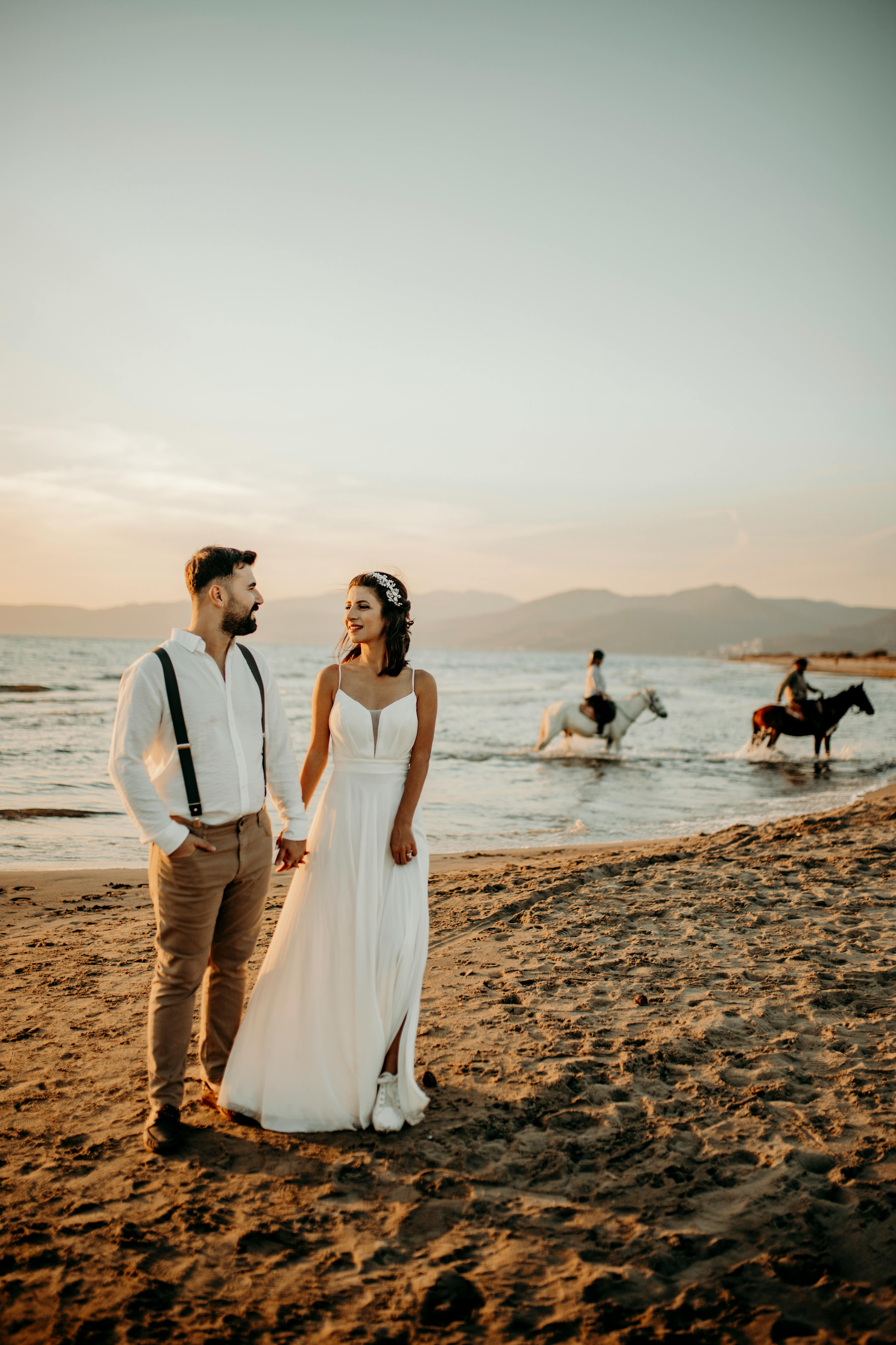 Elegant couple in white attire stands on İzmir beach at sunset, holding hands with horse riders in the background.