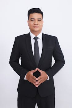 Portrait of a confident adult man in a black suit on a white background, posing with hands clasped.