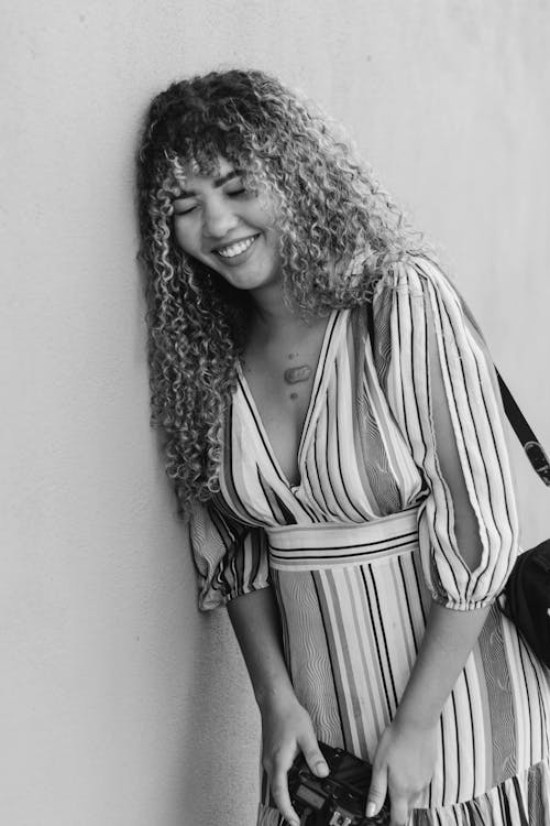 Young Woman with Curly Hair Wearing a Dress Leaning against the Wall and Smiling 
