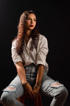 Stylish portrait of a young woman with curly hair and tattoos, sitting on a stool against a black background.