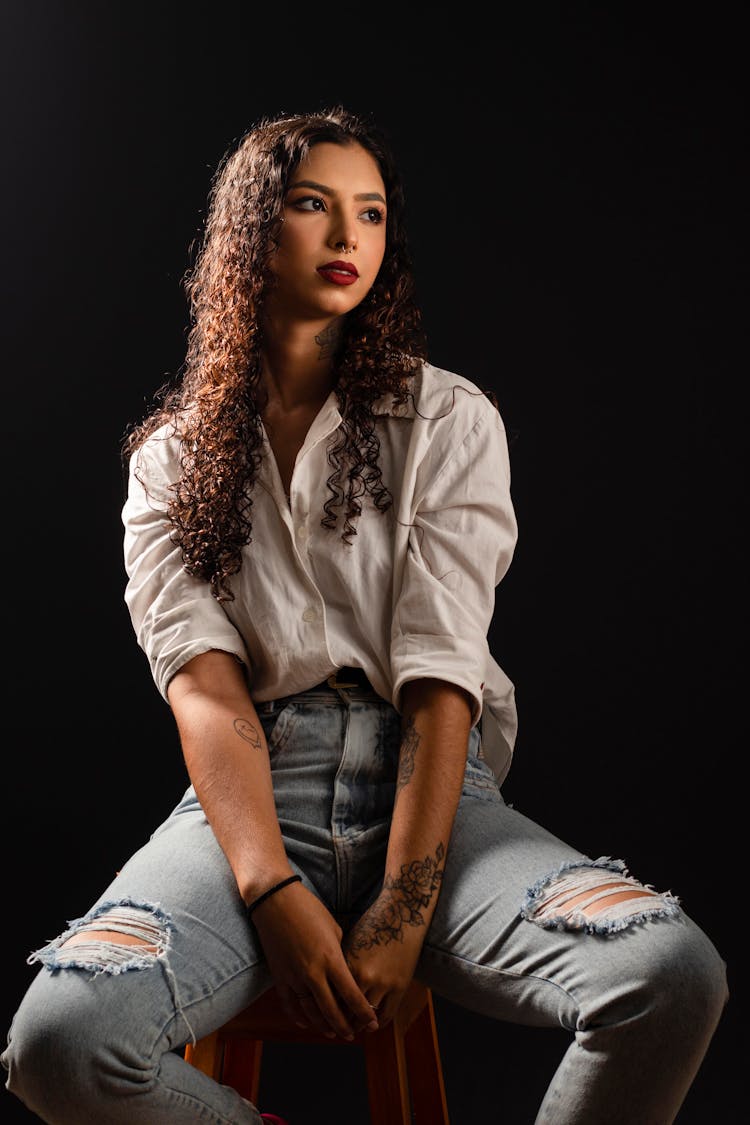 Young Brunette Woman Sitting On A Stool In White Blouse And Light Blue Ripped Jeans