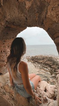 A woman sitting by a stone archway, gazing at the ocean, capturing a moment of relaxation.