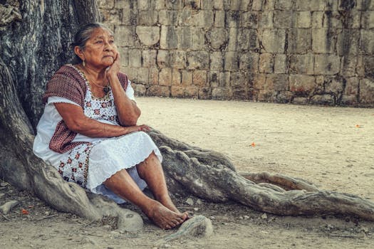An elderly woman sits thoughtfully by a tree at Chichén Itzá, Mexico.