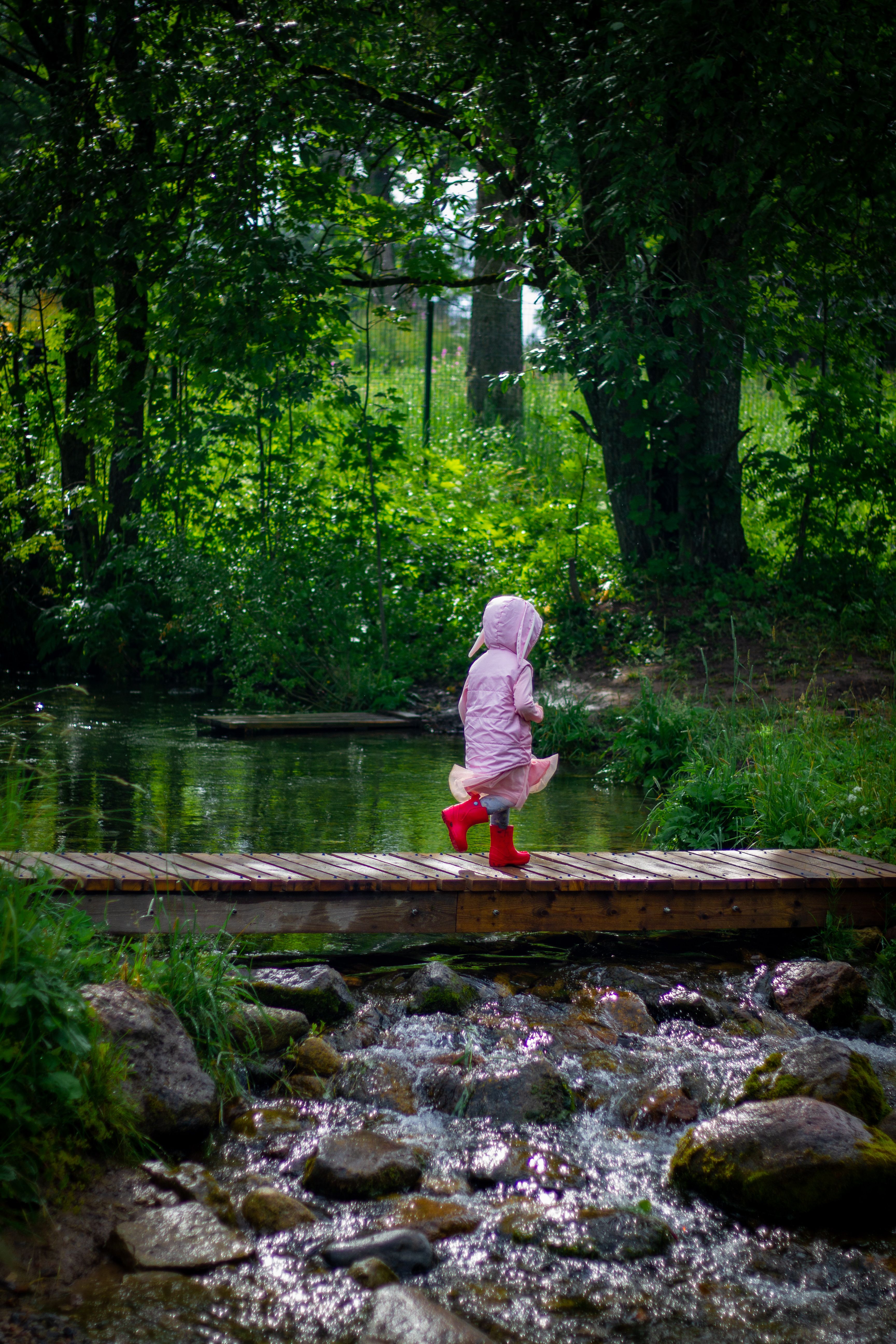 A young child in a pink jacket and red boots runs across a footbridge in a lush green forested park.