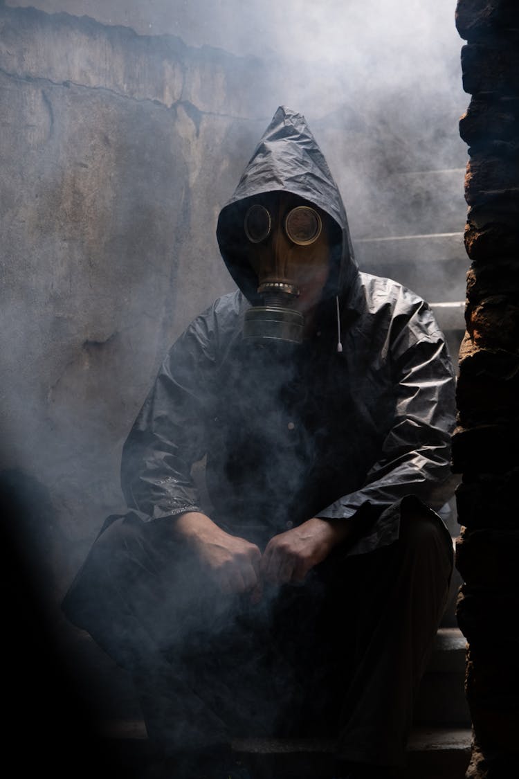 Person In Gas Mask And Protective Suit Sitting On Stairs In A Smoke-filled Abandoned Building